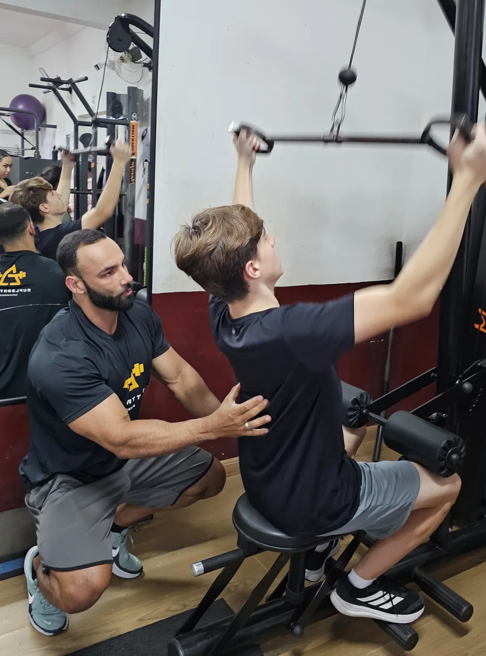 Igor agachado ao lado de um aluno na academia, com a mão no tronco dele corrigindo a postura durante o exercício